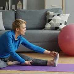 Mujer en azul estirando isquiotibiales en una alfombra morada, con una pelota de ejercicio rosa al lado.