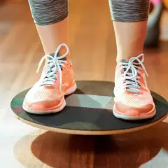 Persona realizando ejercicios de equilibrio sobre una tabla inestable, con zapatillas deportivas naranjas y mallas grises.