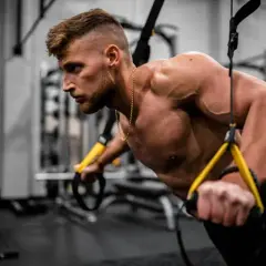 Hombre entrenando en gimnasio, enfocándose en los nombres de los músculos del cuerpo mientras realiza ejercicios de fuerza con TRX.