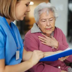 Enfermera revisa notas con una anciana que se lleva la mano al pecho, posiblemente por síntomas de cuerpos de Lewy.