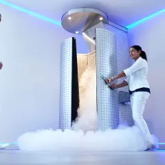 Una mujer en bata blanca se acerca a una cabina de crioterapia que emite vapor. Otra mujer, vestida de blanco, abre la puerta.