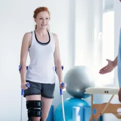 Mujer pelirroja con muletas y rodillera, sonriendo a un fisioterapeuta. Su recuperación avanza con reposo relativo y ejercicios.