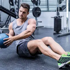 Hombre sentado en el suelo del gimnasio, girando el torso con una pelota medicinal azul, trabajando la elasticidad muscular.
