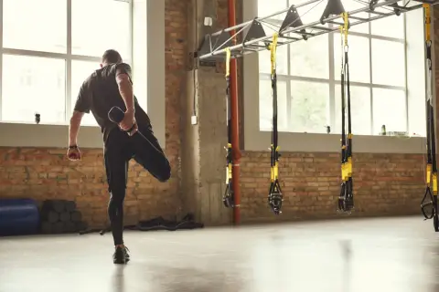 Hombre estirando la pierna, un ejemplo de la definición de calentamiento en un gimnasio con ladrillos y TRX.