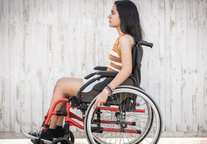Joven en silla de ruedas, con brazaletes, frente a pared texturizada. La ataxia de Friedreich no detiene su espíritu.