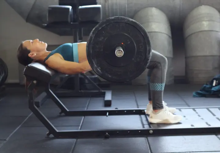 Mujer realizando un hip thrust con barra sobre un banco en un gimnasio.