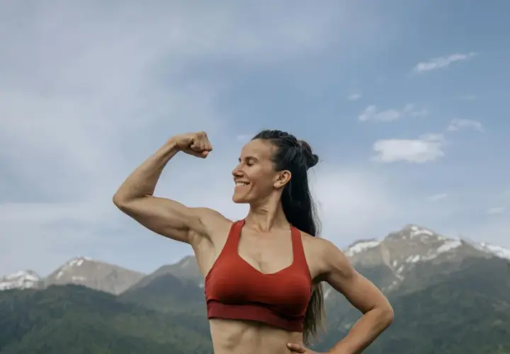 Mujer sonriente muestra su bíceps, demostrando la increíble **función del tejido muscular** en un paisaje montañoso.