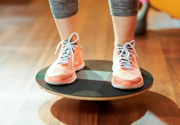 Persona realizando ejercicios de equilibrio sobre una tabla inestable, con zapatillas deportivas naranjas y mallas grises.