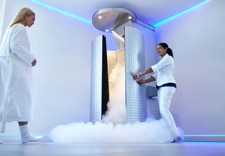 Una mujer en bata blanca se acerca a una cabina de crioterapia que emite vapor. Otra mujer, vestida de blanco, abre la puerta.
