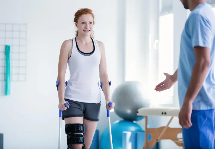 Mujer pelirroja con muletas y rodillera, sonriendo a un fisioterapeuta. Su recuperación avanza con reposo relativo y ejercicios.