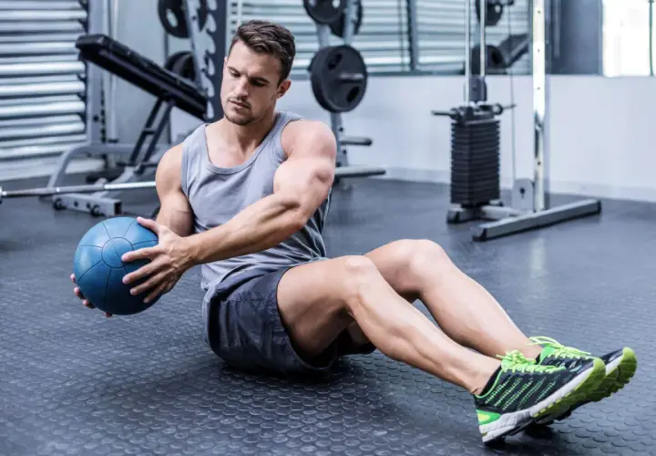 Hombre sentado en el suelo del gimnasio, girando el torso con una pelota medicinal azul, trabajando la elasticidad muscular.