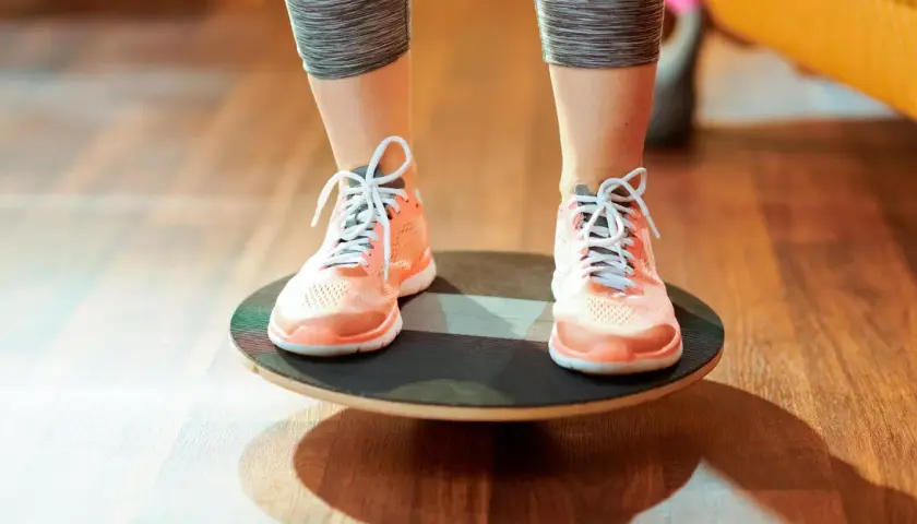 Persona realizando ejercicios de equilibrio sobre una tabla inestable, con zapatillas deportivas naranjas y mallas grises.