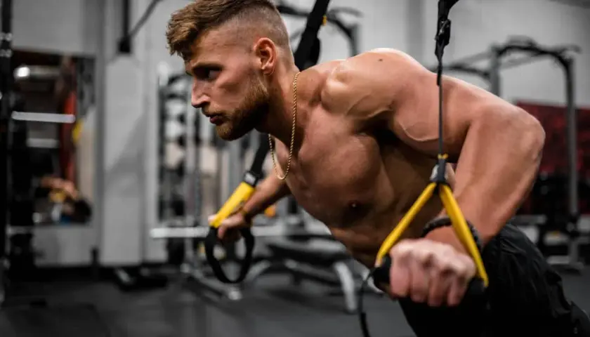 Hombre entrenando en gimnasio, enfocándose en los nombres de los músculos del cuerpo mientras realiza ejercicios de fuerza con TRX.