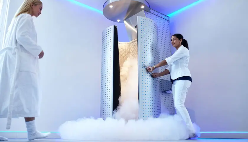 Una mujer en bata blanca se acerca a una cabina de crioterapia que emite vapor. Otra mujer, vestida de blanco, abre la puerta.