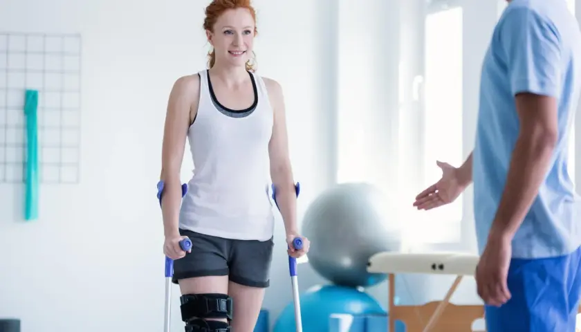 Mujer pelirroja con muletas y rodillera, sonriendo a un fisioterapeuta. Su recuperación avanza con reposo relativo y ejercicios.