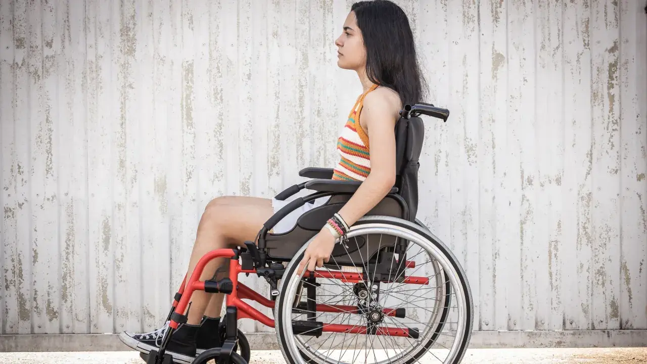 Joven en silla de ruedas, con brazaletes, frente a pared texturizada. La ataxia de Friedreich no detiene su espíritu.