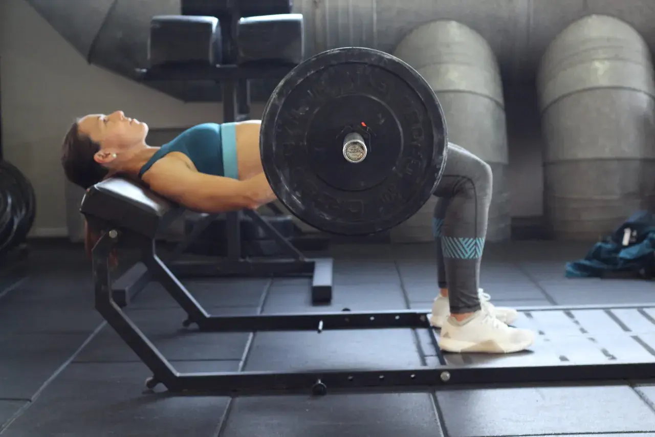 Mujer realizando un hip thrust con barra sobre un banco en un gimnasio.