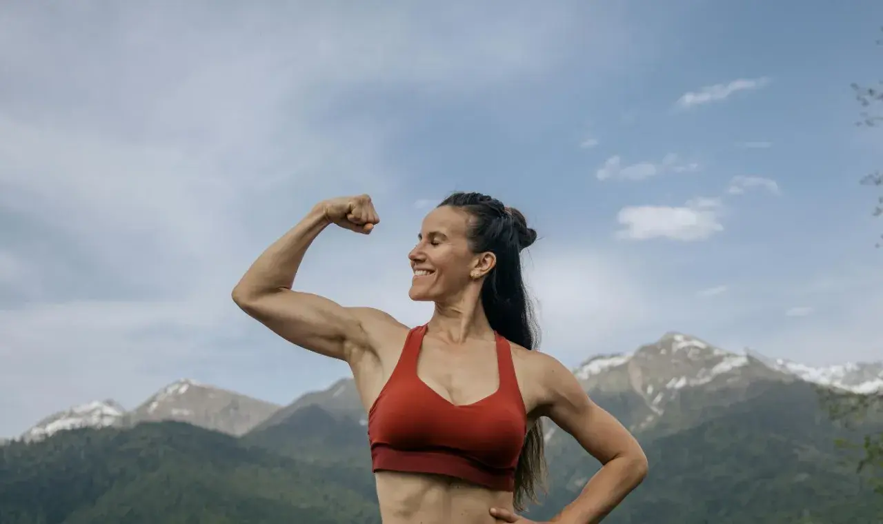Mujer sonriente muestra su bíceps, demostrando la increíble **función del tejido muscular** en un paisaje montañoso.