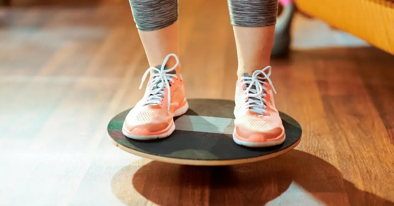 Persona realizando ejercicios de equilibrio sobre una tabla inestable, con zapatillas deportivas naranjas y mallas grises.