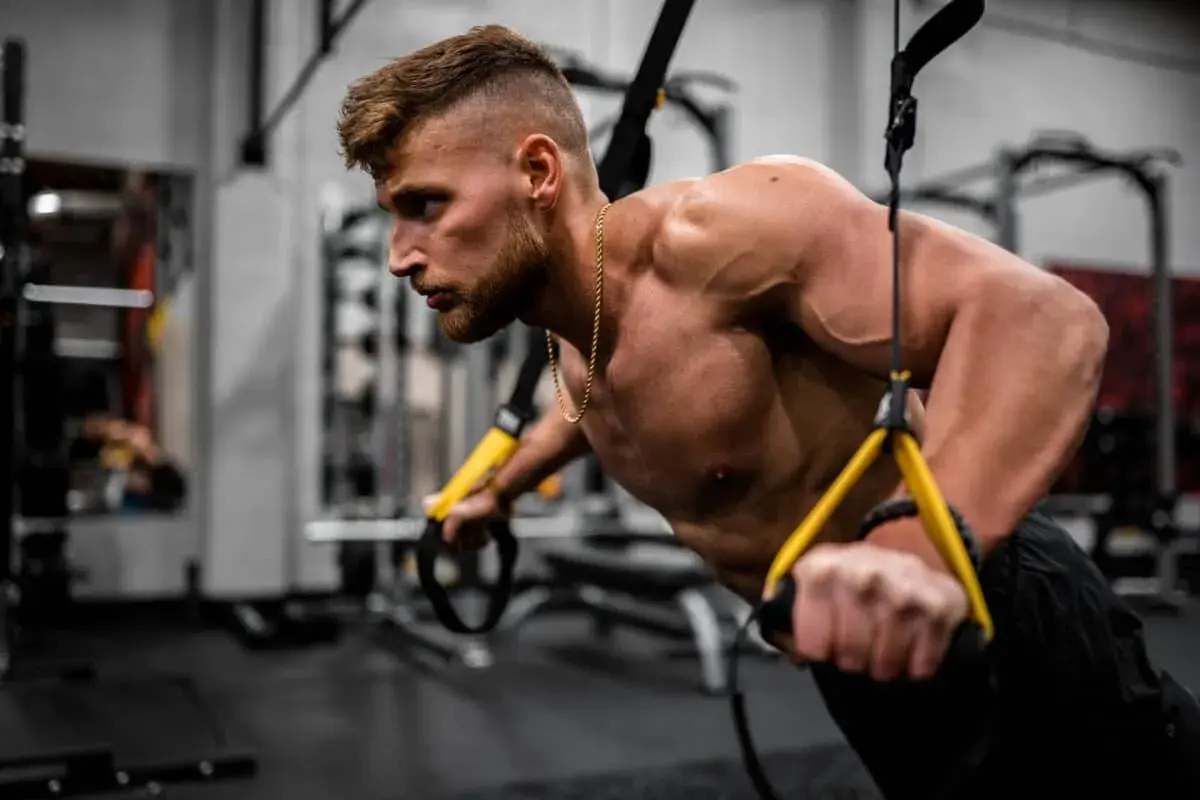 Hombre entrenando en gimnasio, enfocándose en los nombres de los músculos del cuerpo mientras realiza ejercicios de fuerza con TRX.