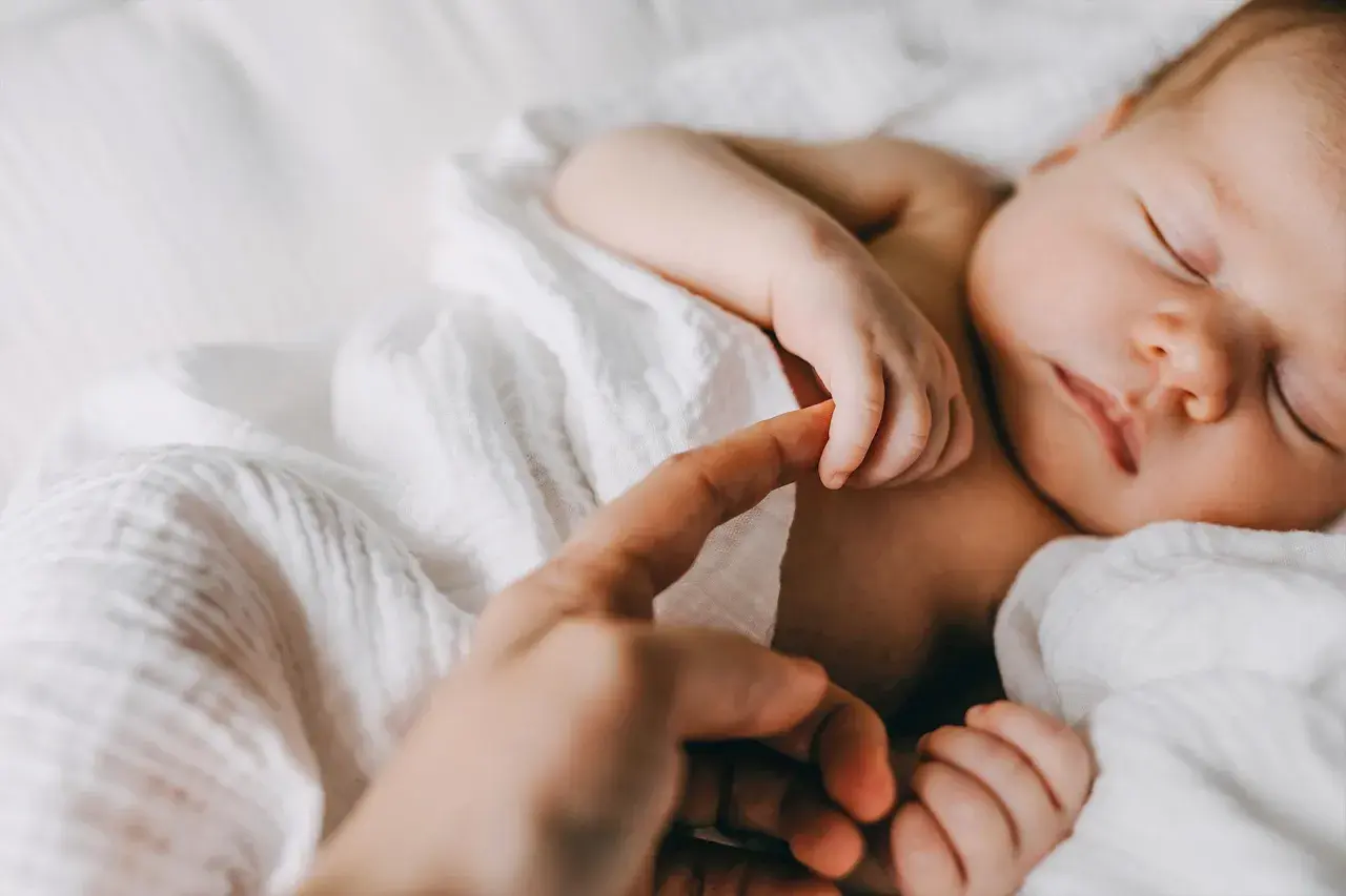 Un bebé duerme plácidamente, su pequeña mano aferra un dedo adulto. Un tierno reflejo de moro en la conexión entre generaciones.