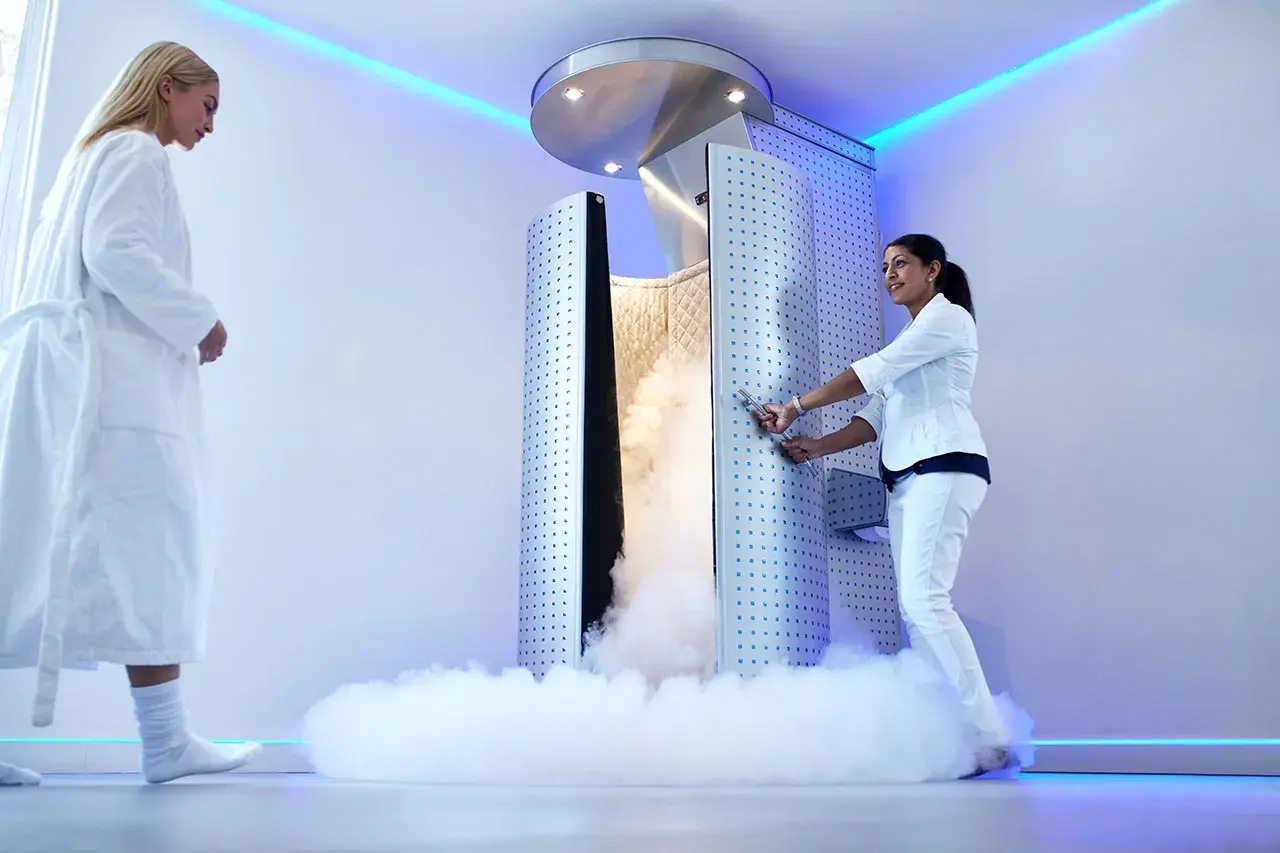 Una mujer en bata blanca se acerca a una cabina de crioterapia que emite vapor. Otra mujer, vestida de blanco, abre la puerta.