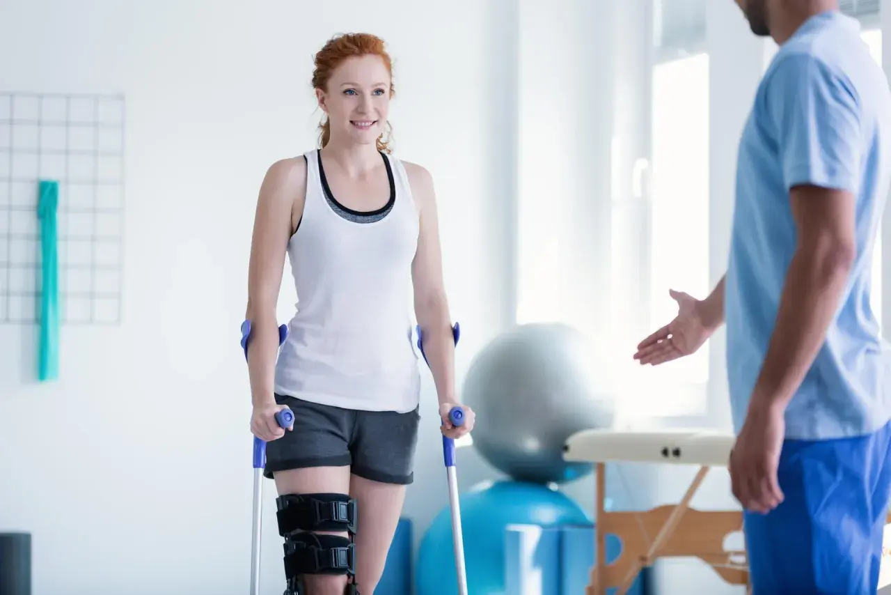 Mujer pelirroja con muletas y rodillera, sonriendo a un fisioterapeuta. Su recuperación avanza con reposo relativo y ejercicios.