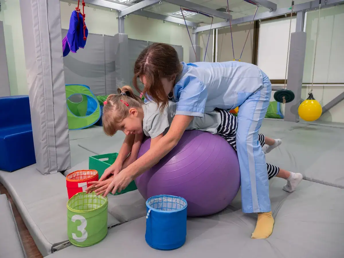 Niña con parálisis cerebral se ejercita en una pelota de fisioterapia, guiada por un terapeuta.