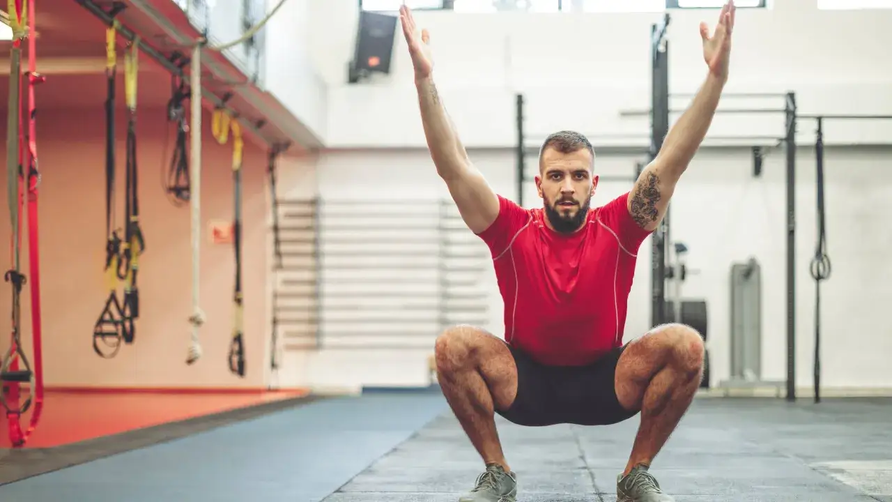 Hombre en gimnasio haciendo sentadilla, buscando la hipertrofia muscular. Brazos arriba, concentración total.