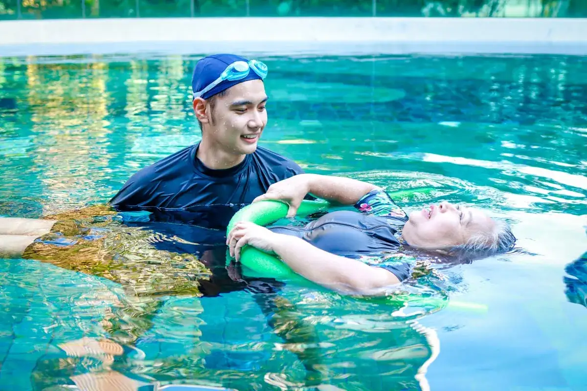 Hombre sonriente ayuda a mujer a flotar con un flotador verde, disfrutando de los beneficios de la hidroterapia en una piscina.