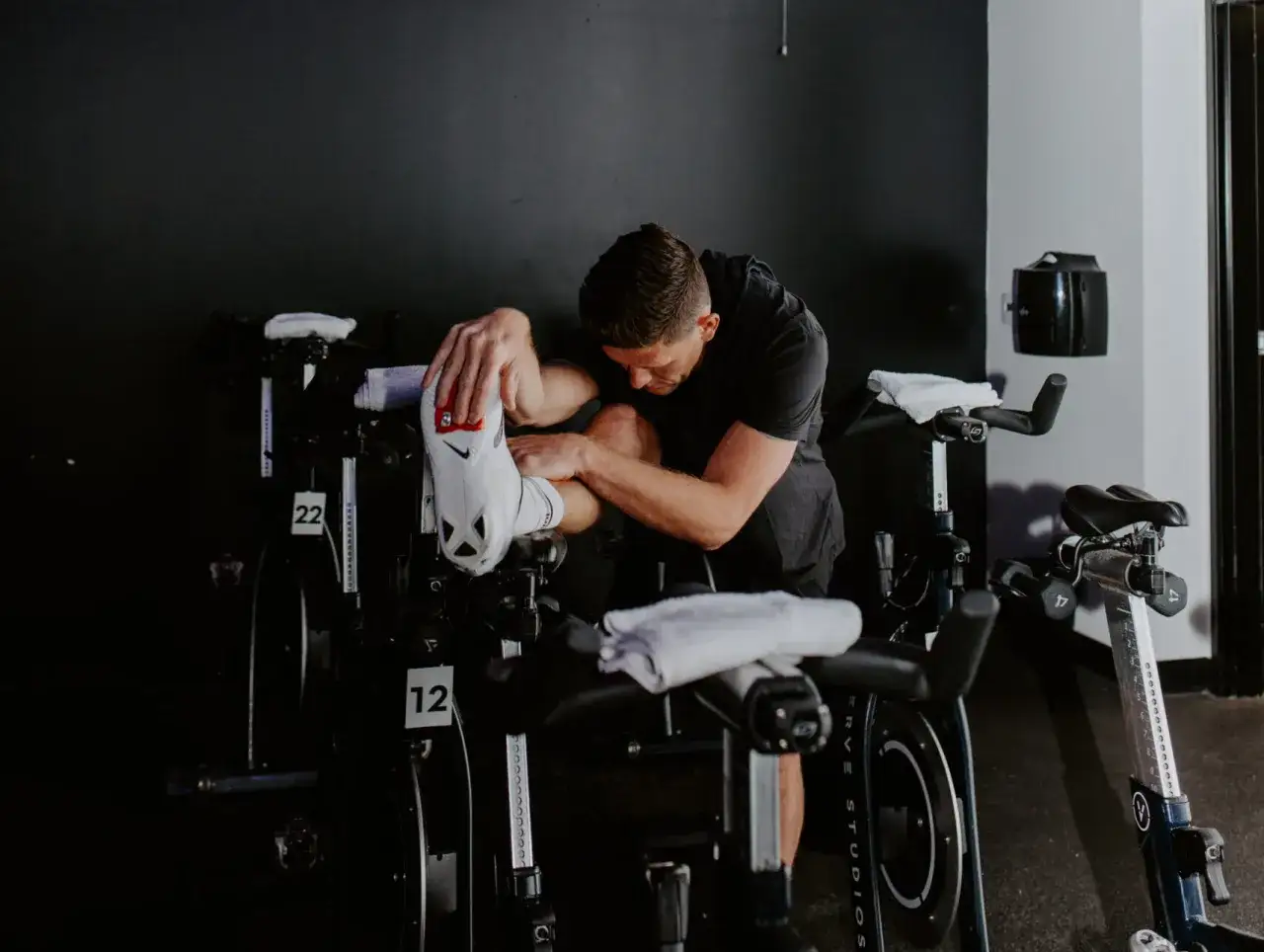 Hombre realizando estiramientos de ciclismo, sentado en una bicicleta est&aacute;tica, con la pierna levantada y sujetando el pie.