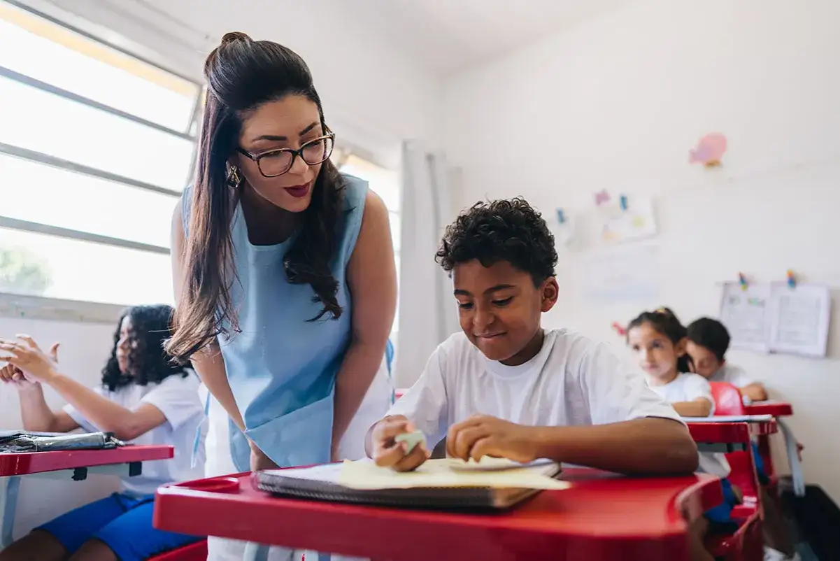 Maestra ayuda a un niño con síndrome de Down en clase. Ambos sonríen mientras trabajan en sus cuadernos.