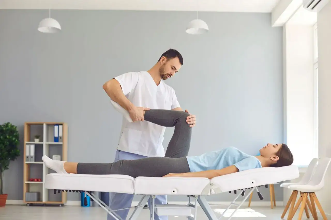Fisioterapeuta ayuda a paciente con estiramiento de pierna en camilla. Un servicio de fisioterapia domiciliaria para tu recuperaci&oacute;n.